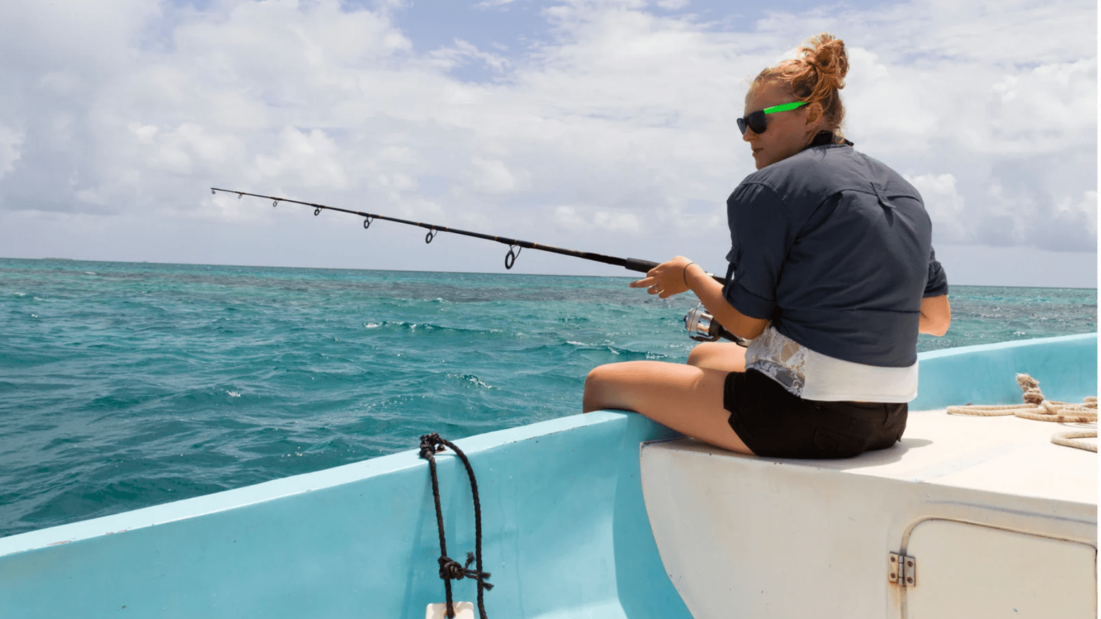 A person in sunglasses is fishing from a boat in clear blue water under a cloudy sky.