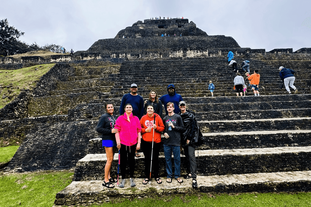 A group of people stands smiling in front of a large ancient stone pyramid with wide steps. Visitors are climbing the structure under cloudy skies.