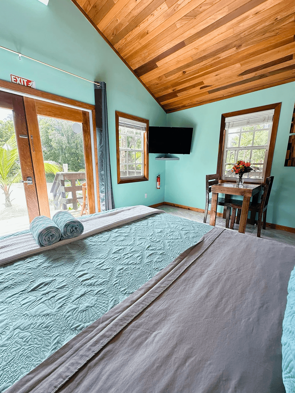 Cozy bedroom with a turquoise wall, wooden ceiling, a TV, a dining table, and doors leading outside.