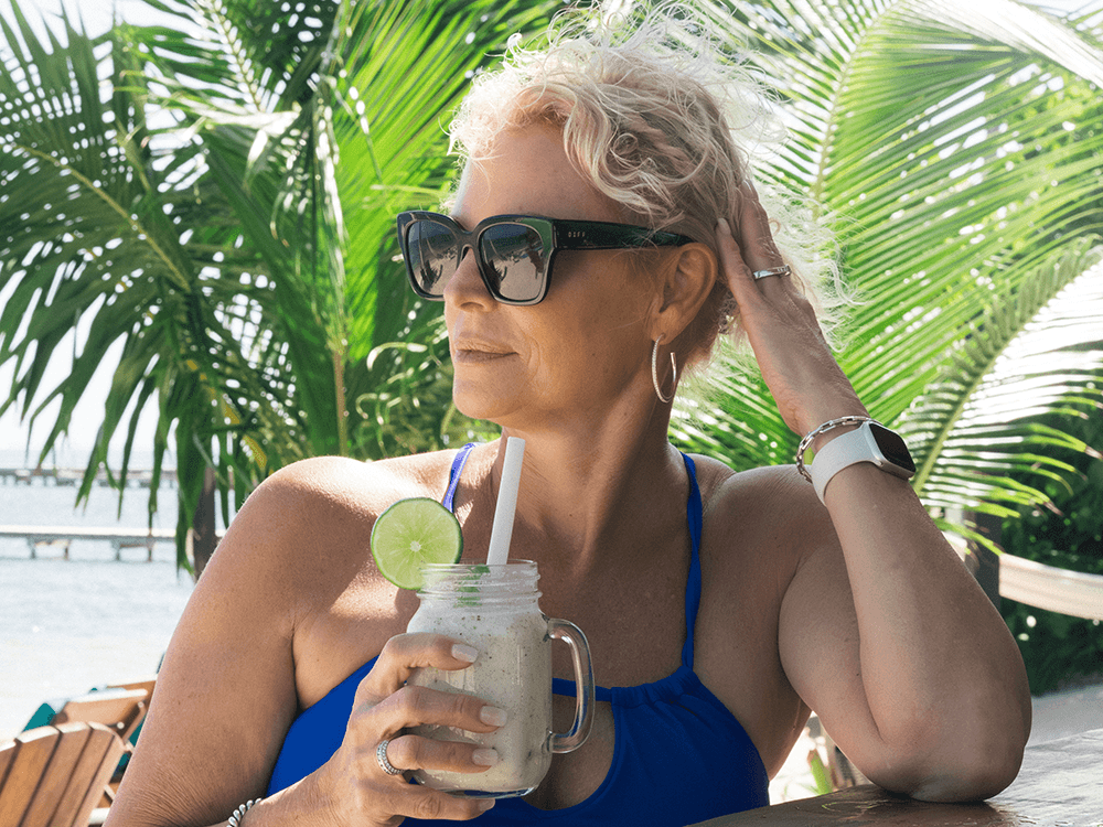 A woman in a blue swimsuit sips a drink with a lime slice while relaxing near palm trees.
