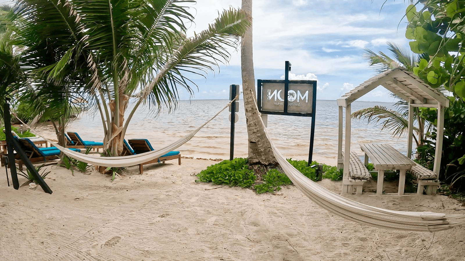 A serene beach scene featuring two hammocks between palm trees, beach chairs, and a small hut near the water.