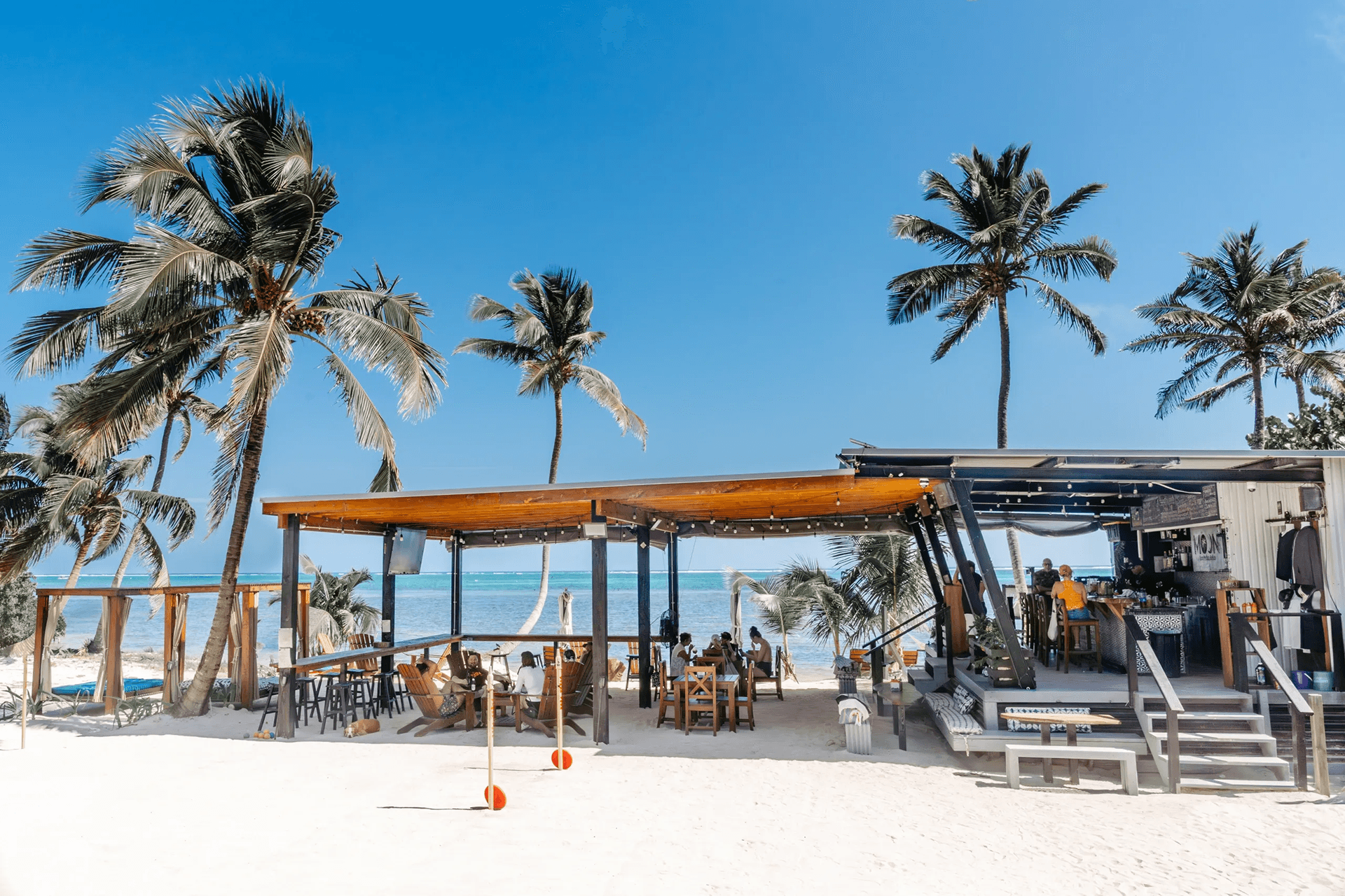 A beachside restaurant surrounded by palm trees and a clear blue ocean.