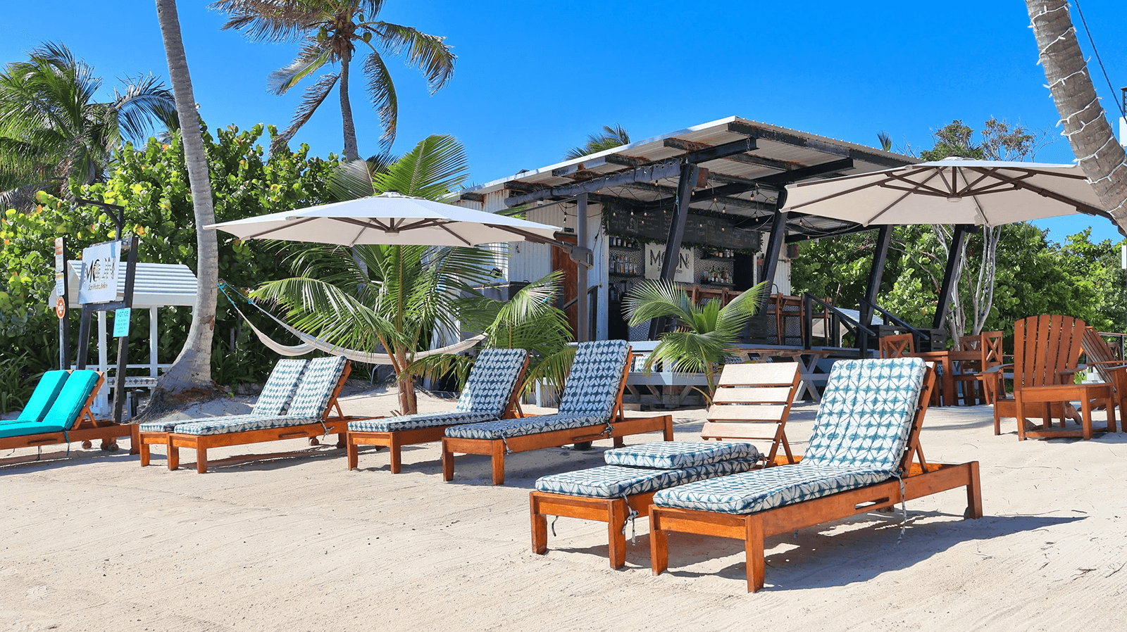 Beachside lounge chairs under umbrellas with a bar and palm trees in the background.