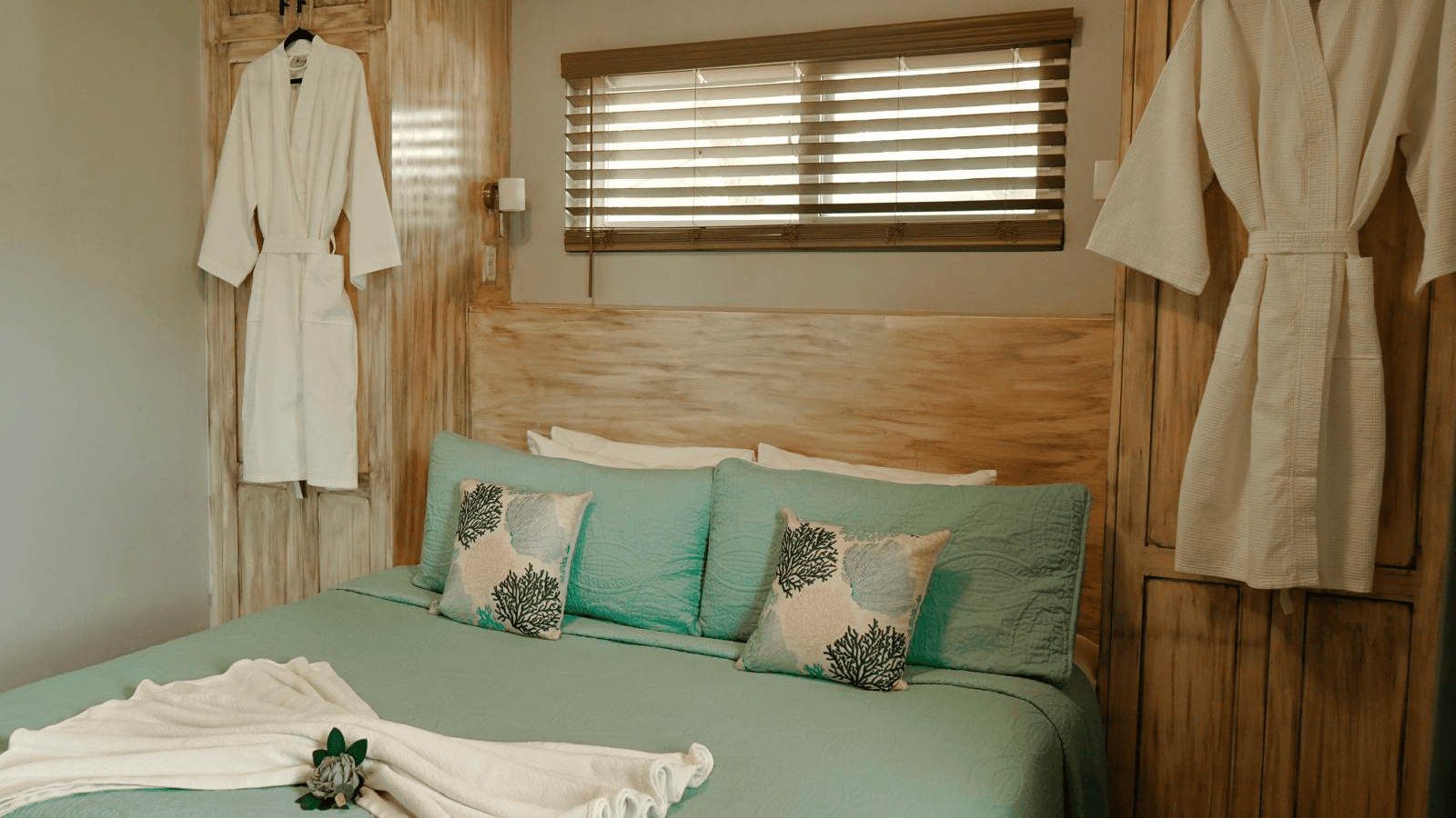 A cozy bedroom featuring a turquoise bedspread, decorative pillows, and bathrobes hanging on the wall.