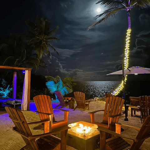 Cozy beachside setting with wooden chairs around a fire pit, illuminated by moonlight and soft lights.