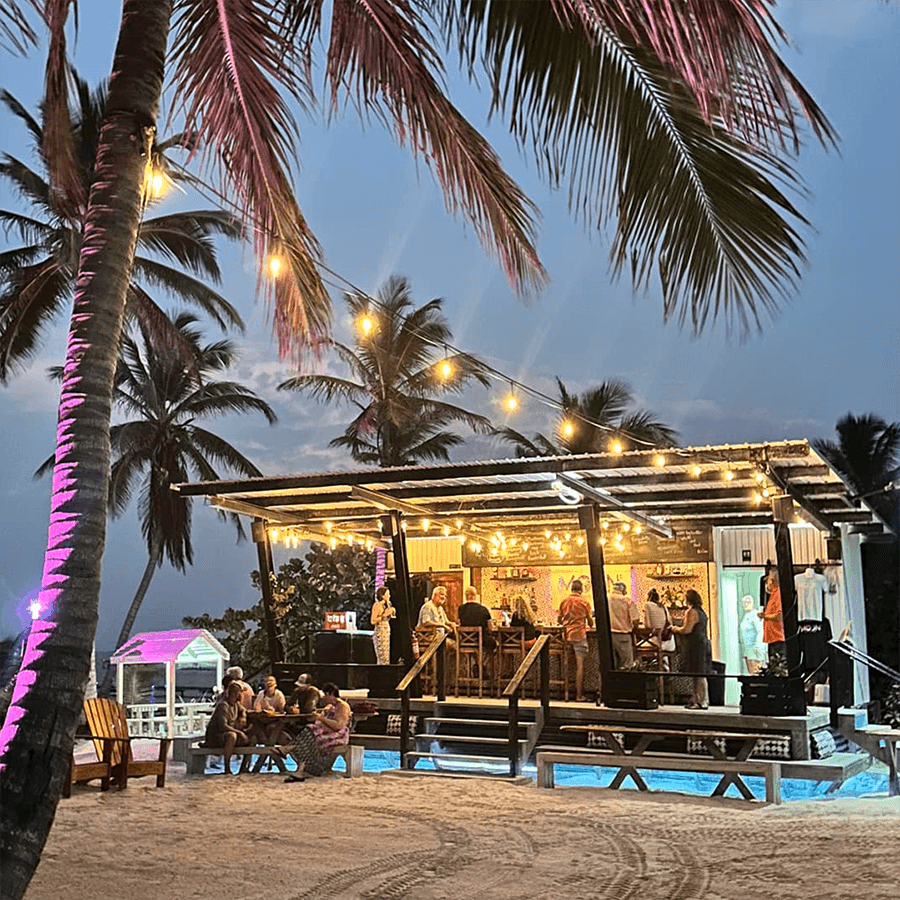 A lively beach bar with palm trees and string lights, featuring people socializing as night falls.