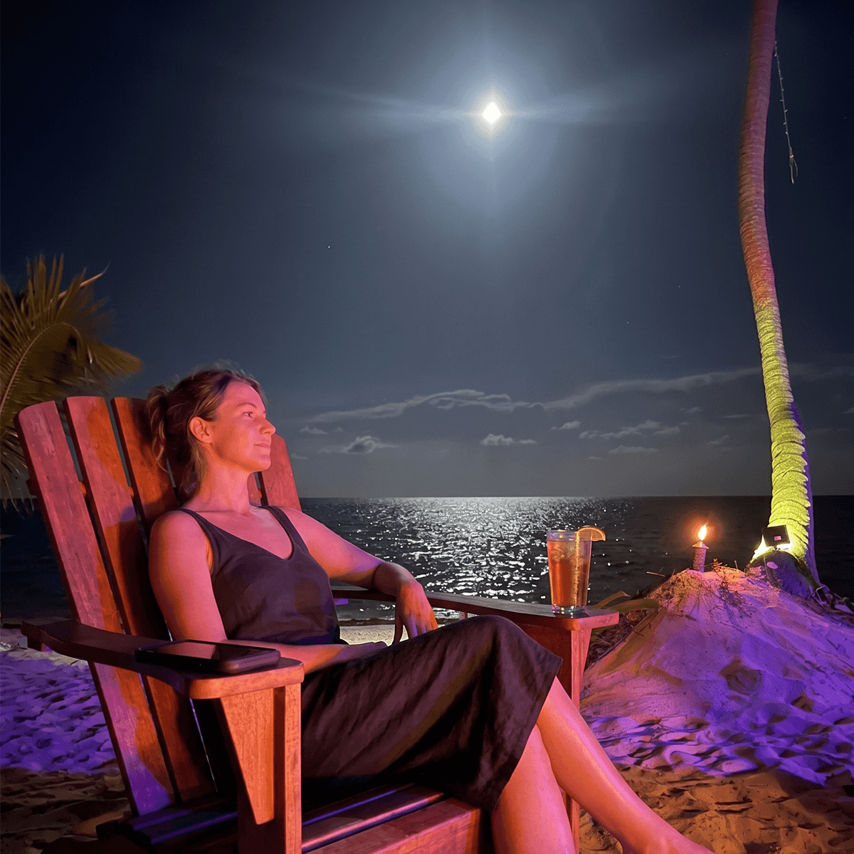 A woman relaxes in a beach chair by the moonlit sea, holding a drink with a candle nearby.