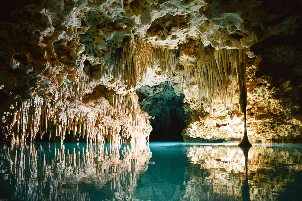 A serene underground cave with stalactites reflecting in calm blue water.