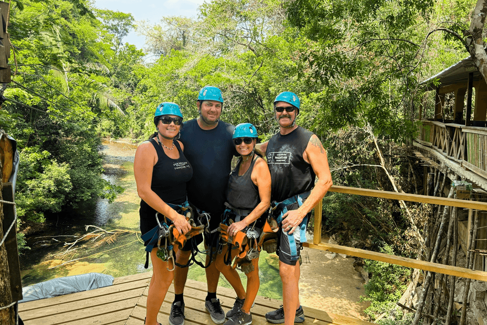 Four people in helmets and harnesses smile on a zipline platform surrounded by lush greenery and a river. The scene conveys excitement and adventure.