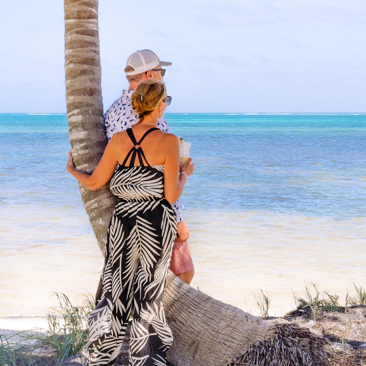 A couple stands together by the shore, leaning against a palm tree with a serene ocean view.