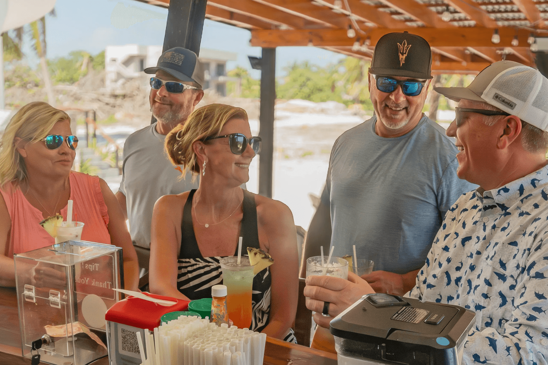 A group of five friends enjoys drinks at a beach bar under sunny skies.