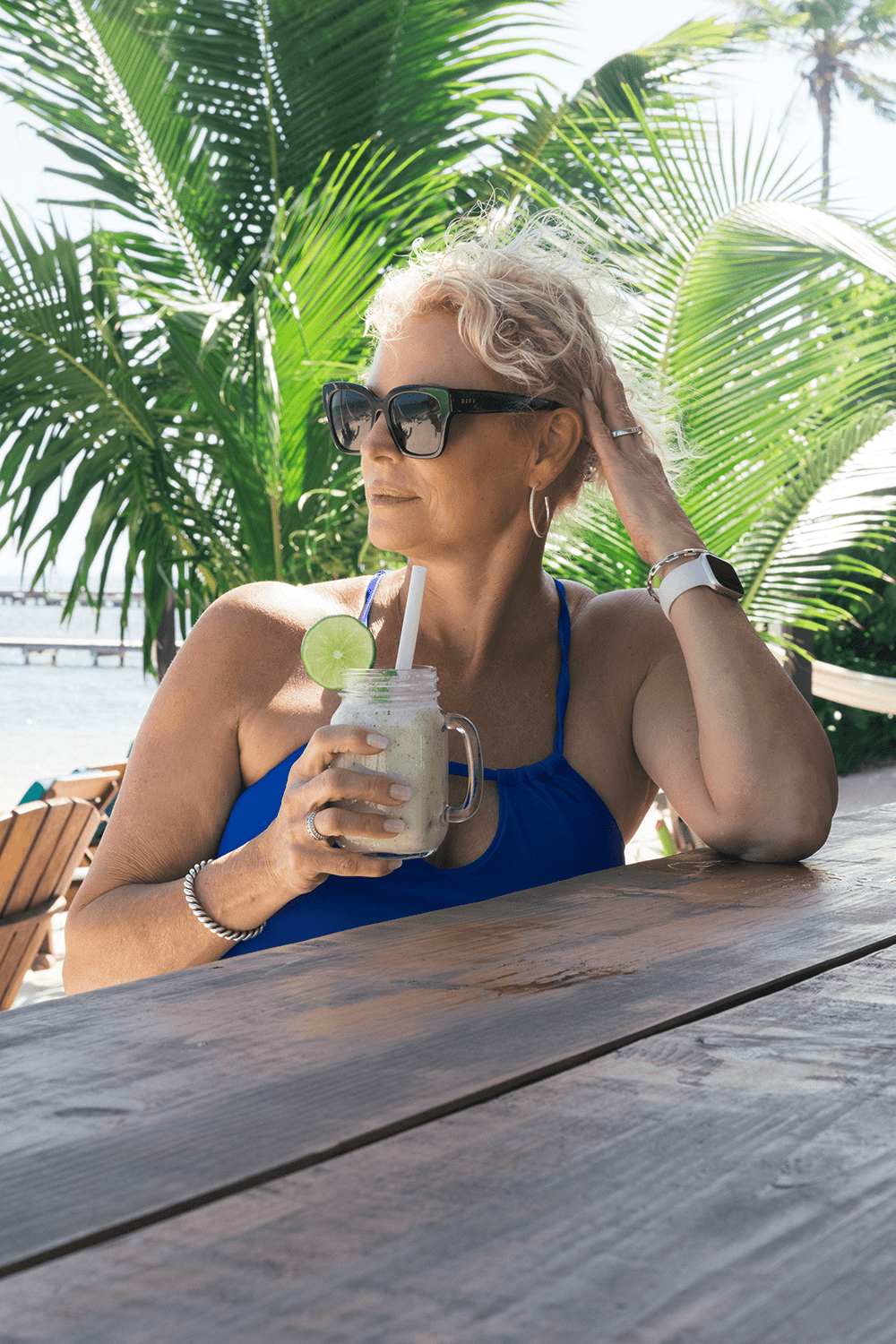 A woman in sunglasses enjoys a tropical drink while sitting at a wooden table surrounded by palm trees.