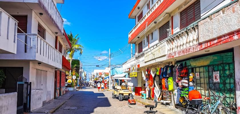 A street view of San Pedro town in Belize with a parked golf cart and two people walking past colorful storefronts.
